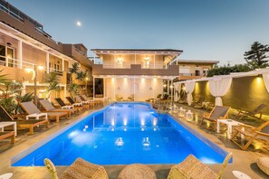 A swimming pool with lounge chairs and umbrellas, surrounded by a building with multiple floors and balconies. - Anemos Suites by Estia (Hersonissos)