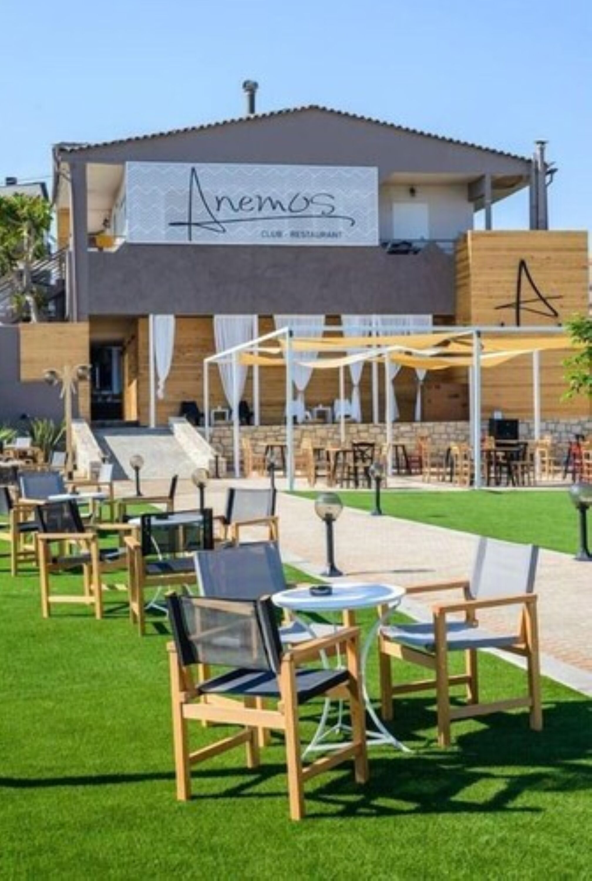 A modern building with a sign reading "Anemos" and outdoor seating with tables and chairs.
