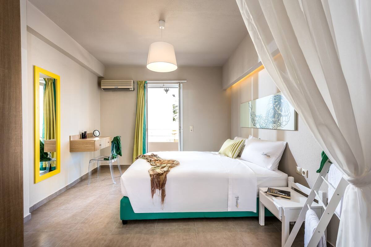 Una habitación de hotel moderna con una cama grande, una mesita de noche, una silla, un espejo y una ventana con cortinas.