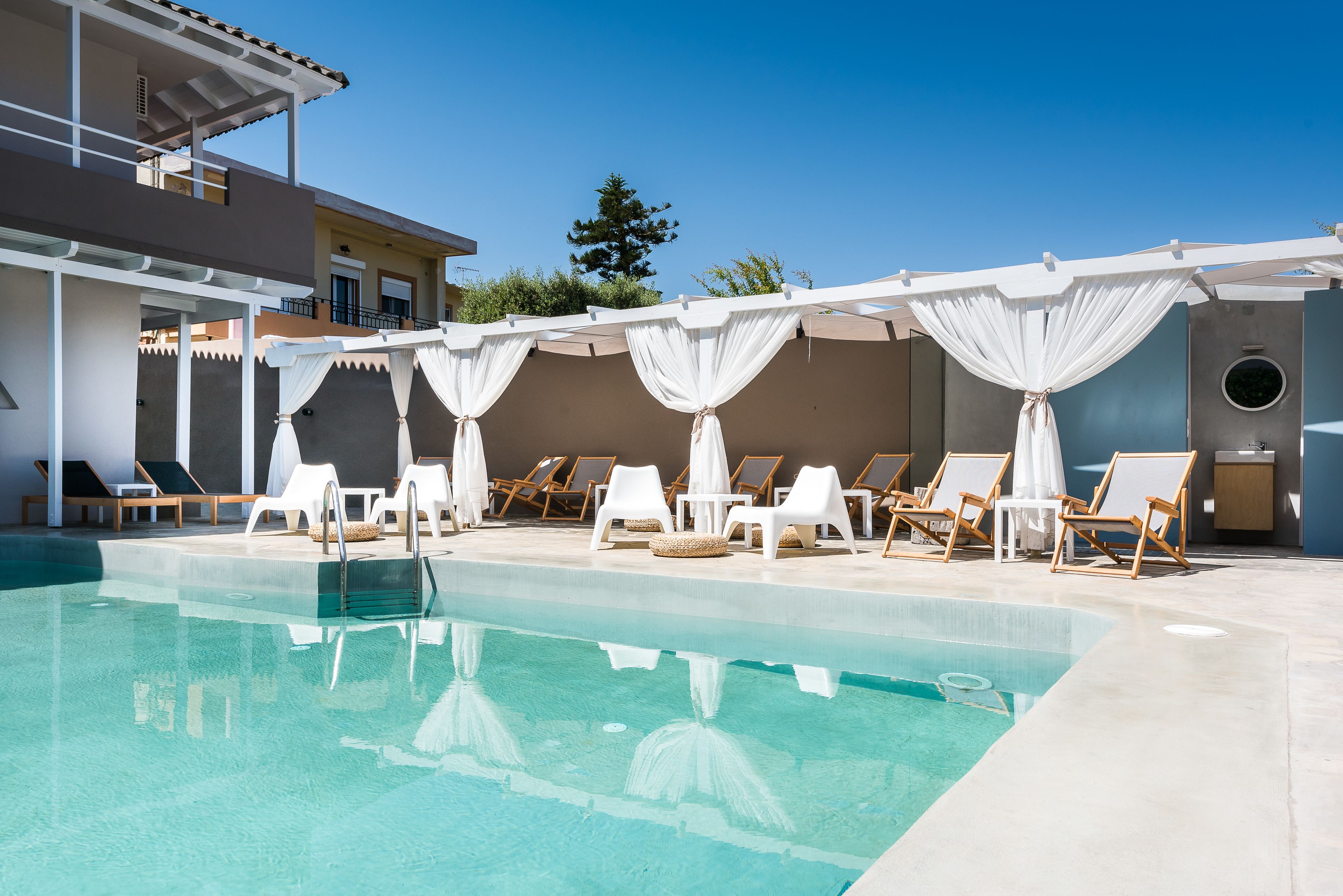 A swimming pool with white curtains, sun loungers, and a building with a balcony.