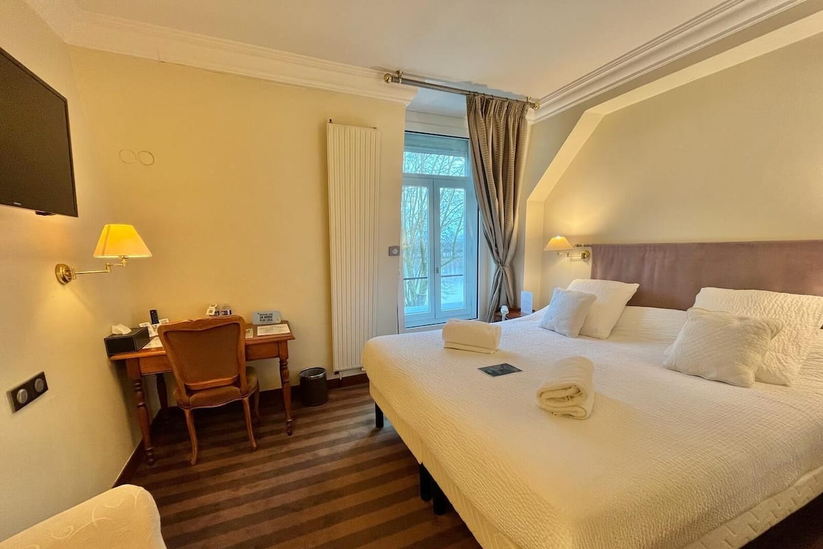 A hotel room with a large bed, a desk with a chair, a TV mounted on the wall, and a window with curtains.