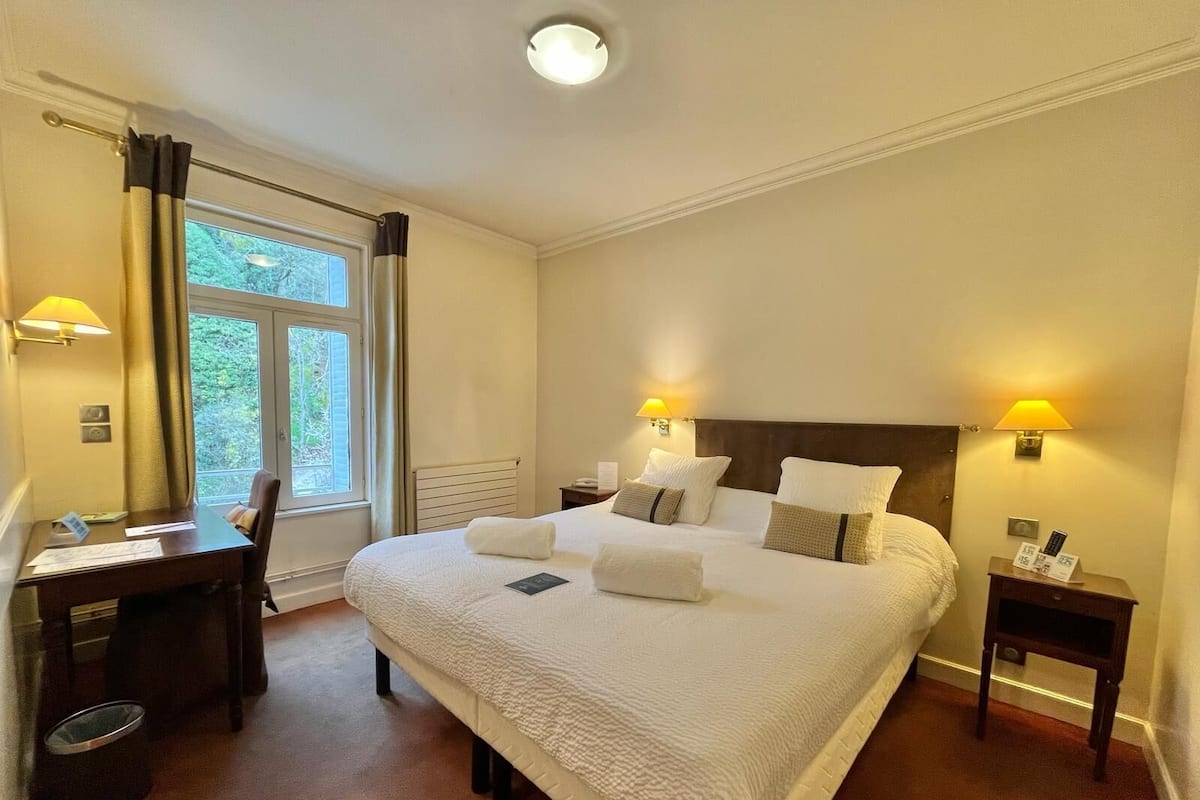 A hotel room with a large bed, two bedside lamps, a desk with a chair, and a window with curtains.