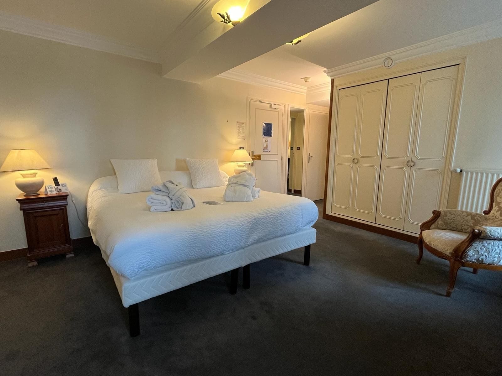Una habitación de hotel con una cama grande, dos lámparas de noche, una mesita de noche de madera, un armario y una silla.