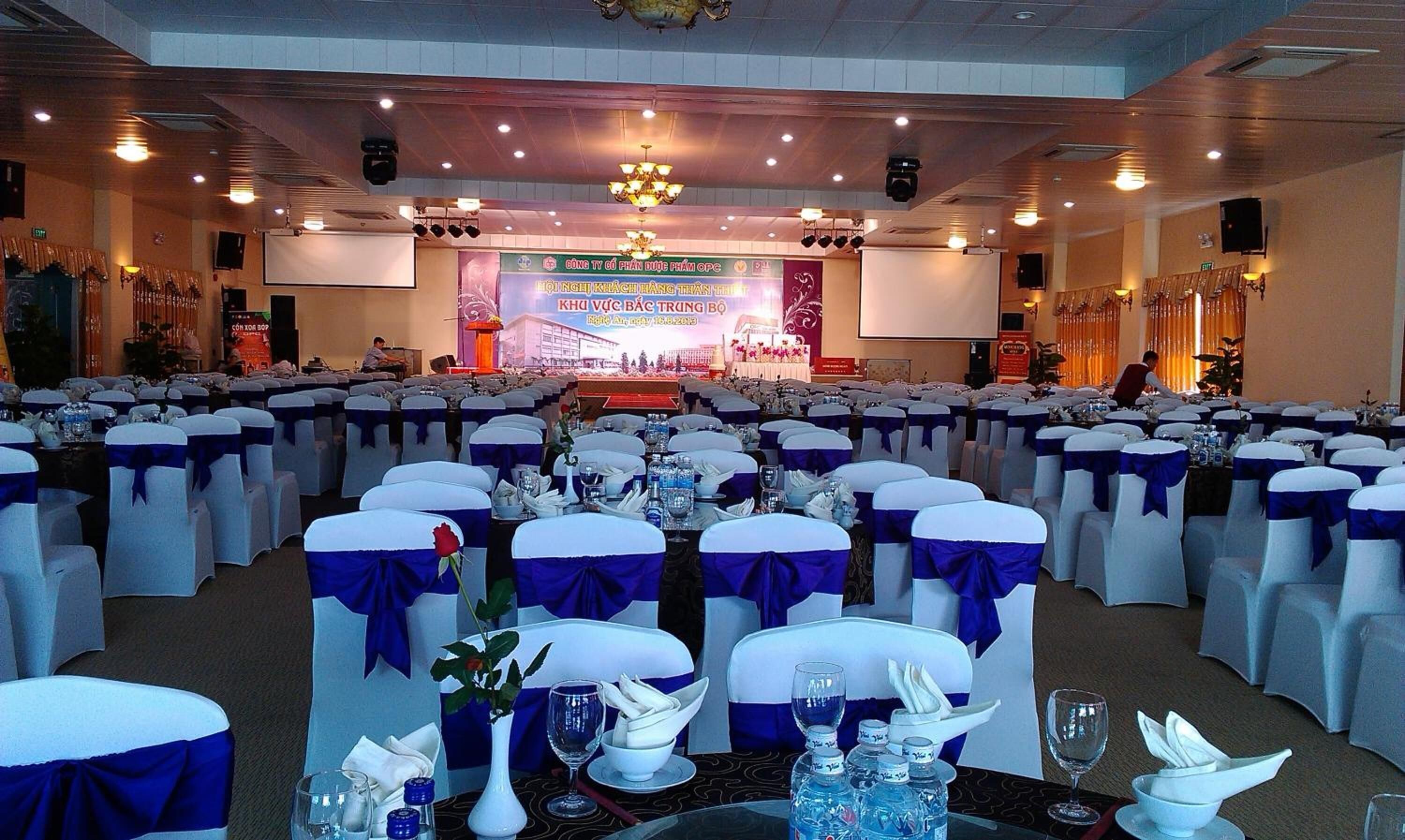 Un salón de banquetes con filas de mesas dispuestas para un evento formal, con sillas blancas adornadas con cintas azules y mesas con botellas de agua y vasos.