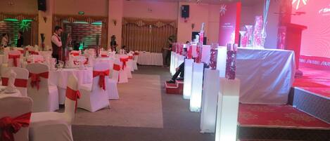Un salón de banquetes con mesas y sillas blancas, cintas rojas y un escenario con alfombra roja.