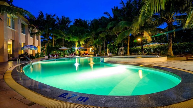 Una piscina con un deck circundante, palmeras y un edificio con mesas al aire libre.