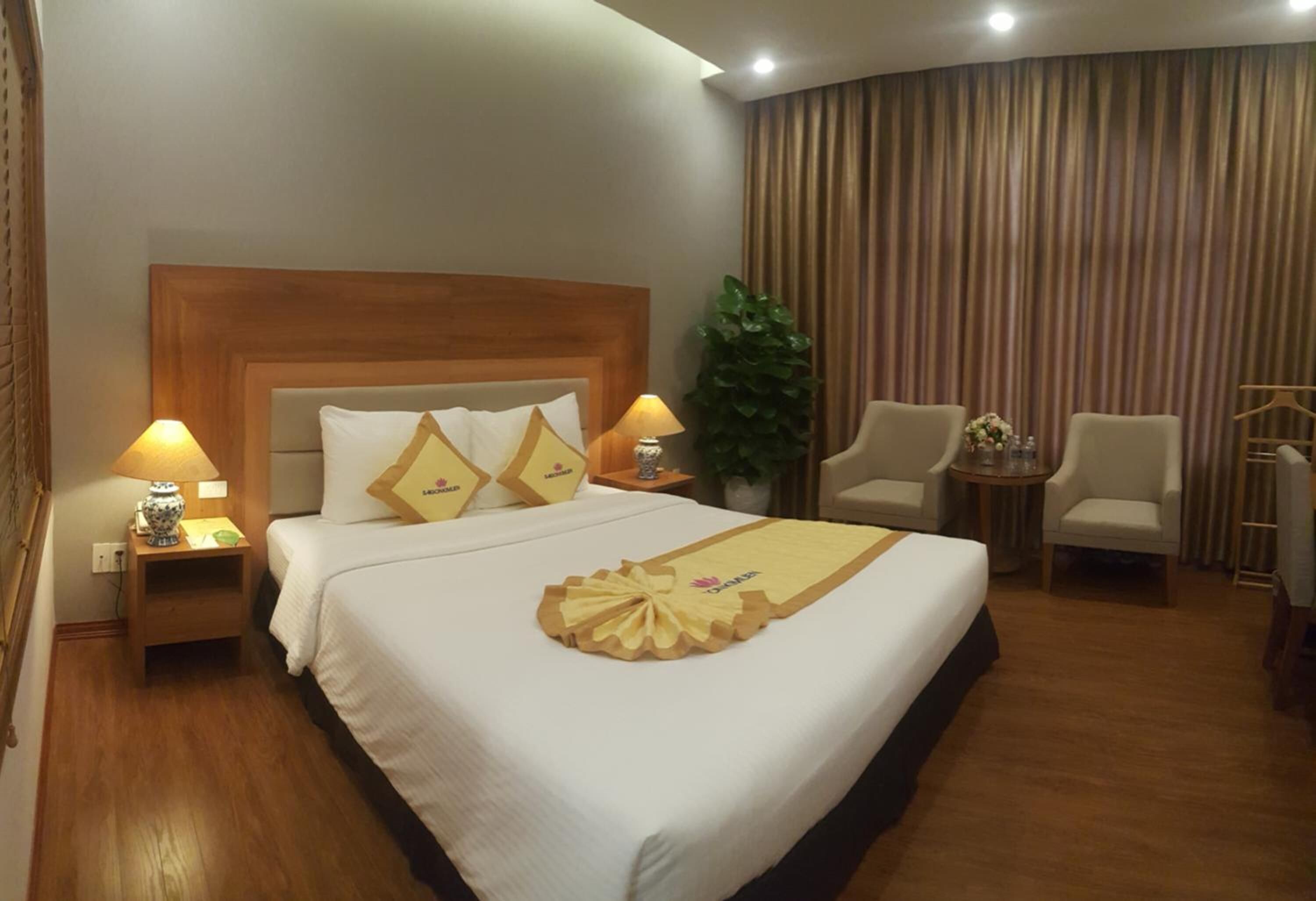 A hotel room with a large bed, two bedside tables with lamps, a small table with a vase, two chairs, and a curtain.