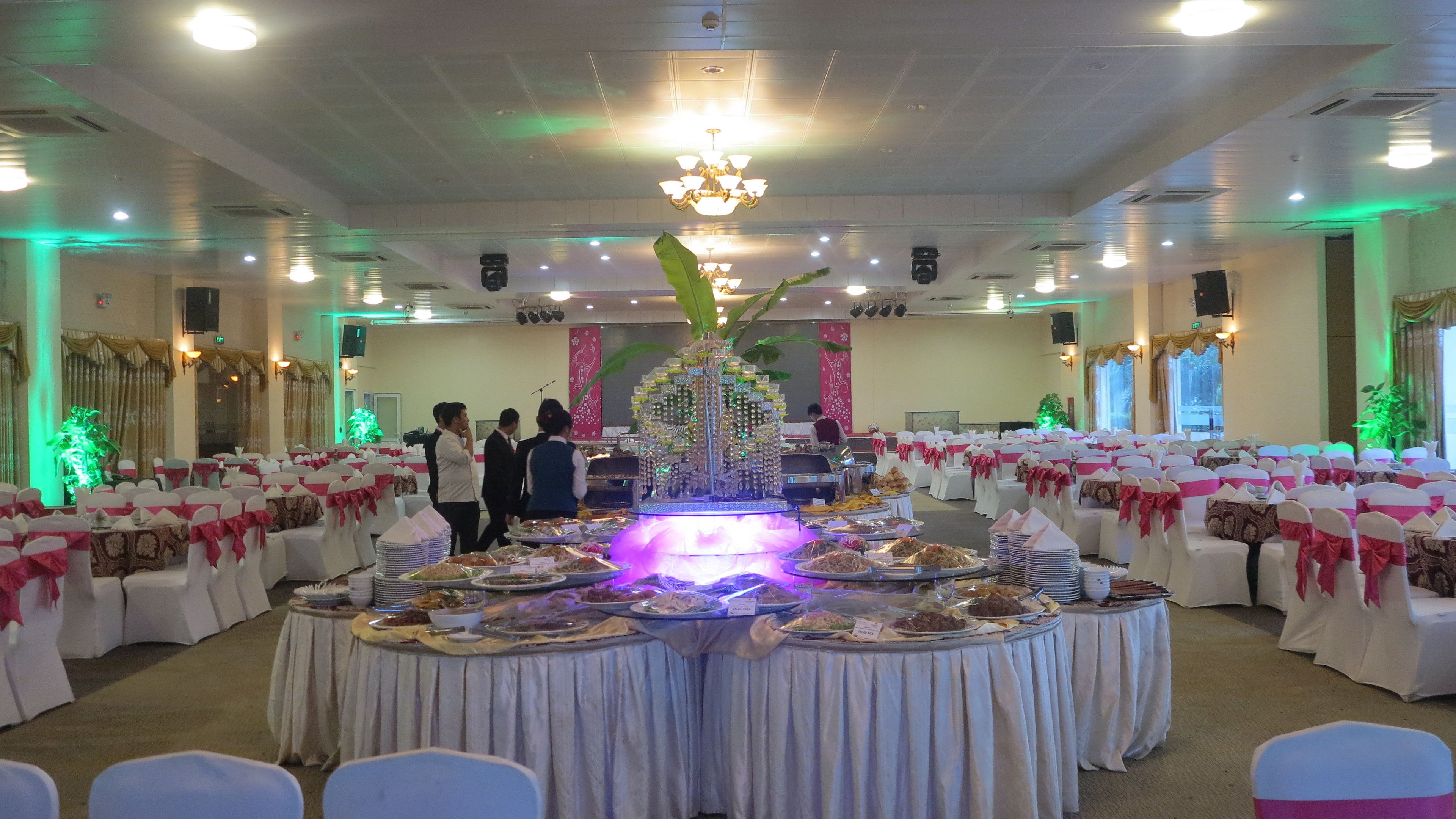 A banquet hall with round tables covered in white tablecloths, surrounded by white chairs with pink sashes. A large decorative centerpiece is in the center.