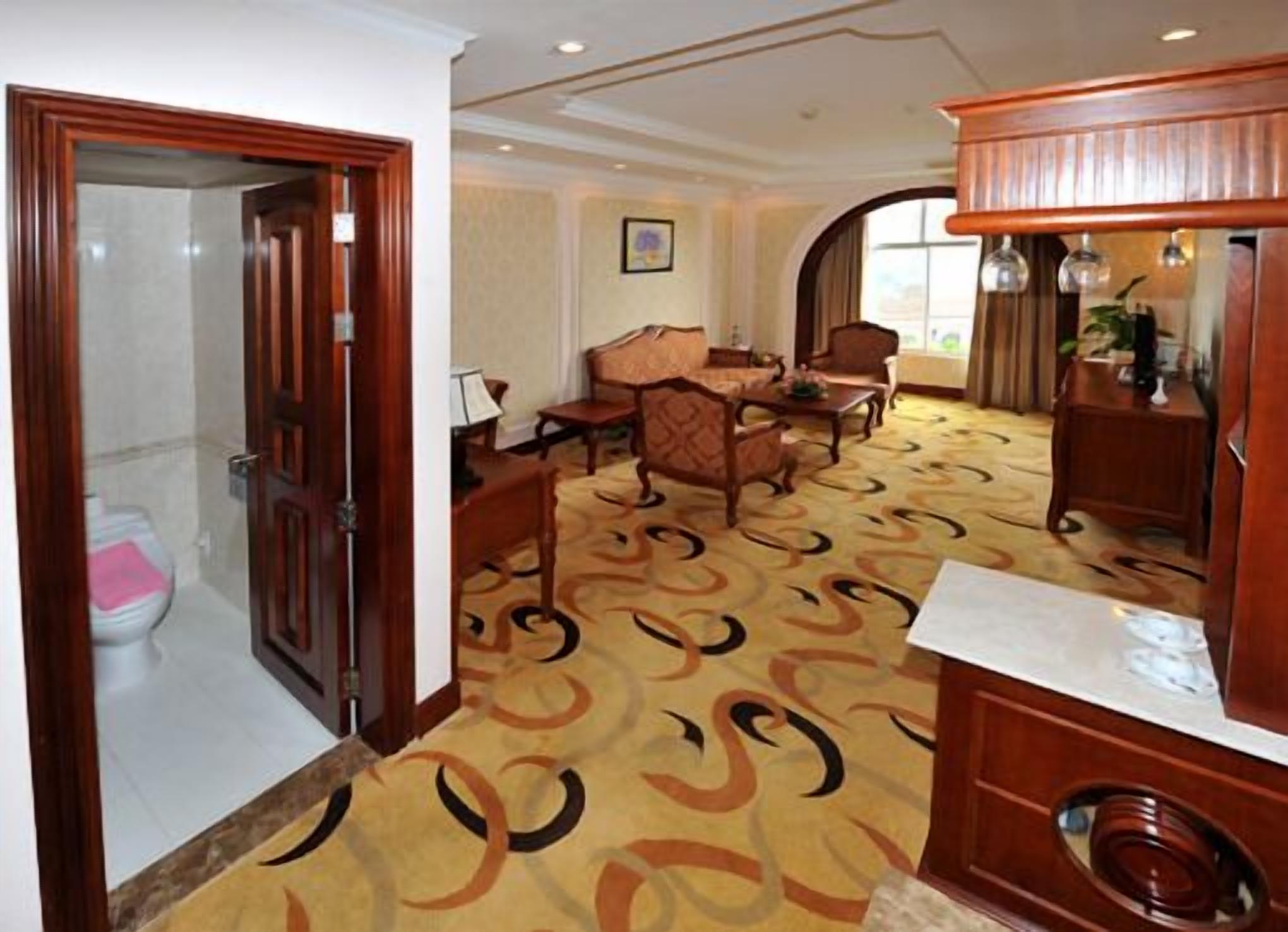 Een hotelkamer met een houten deur, een badkamer met toilet, een zithoek met tafel en stoelen, en een kamerbreed tapijt met dessin.