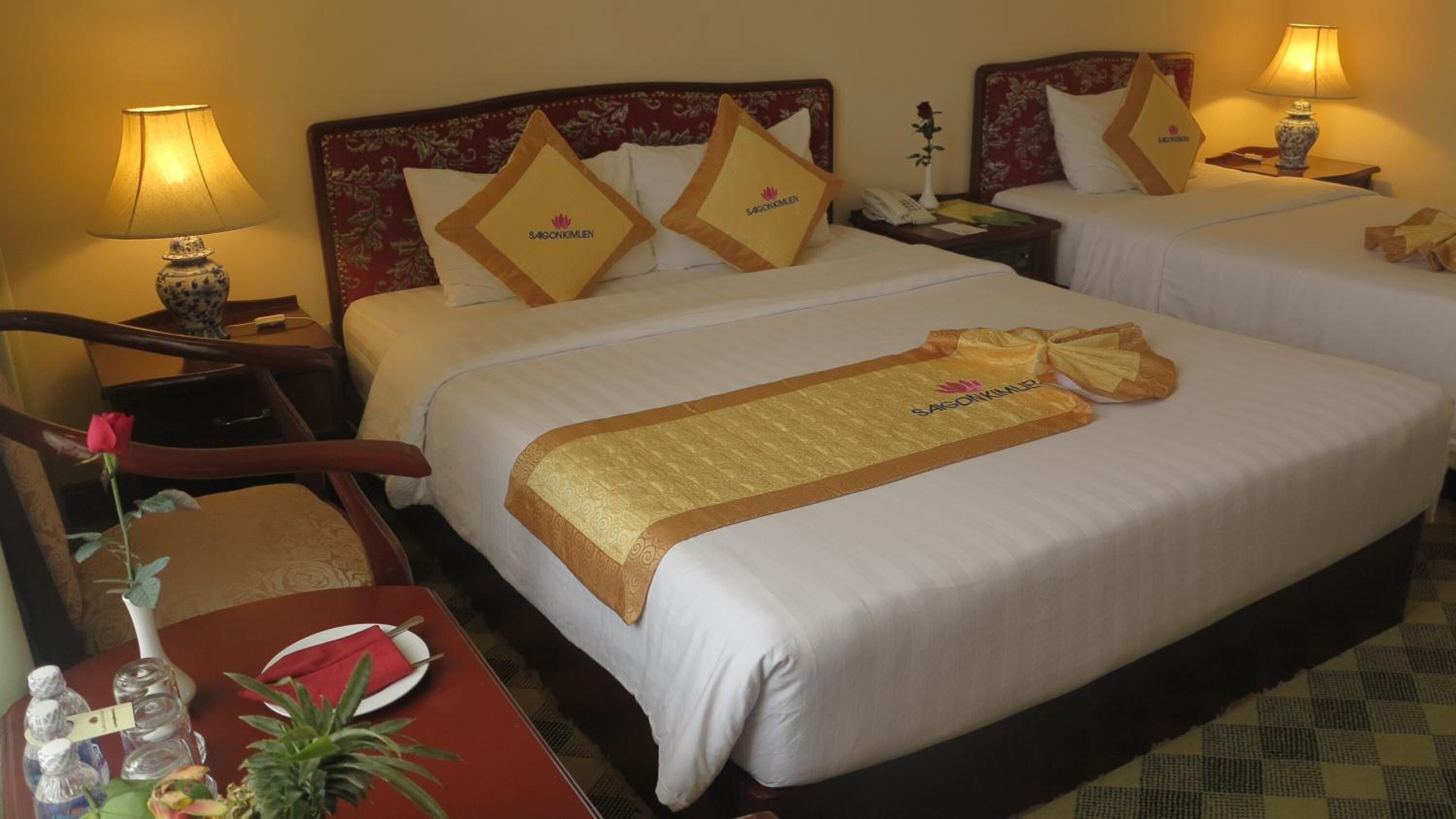 A hotel room with two beds, each with a floral headboard and matching pillows. There are bedside tables with lamps, a small table with a rose and a vase, and a chair.