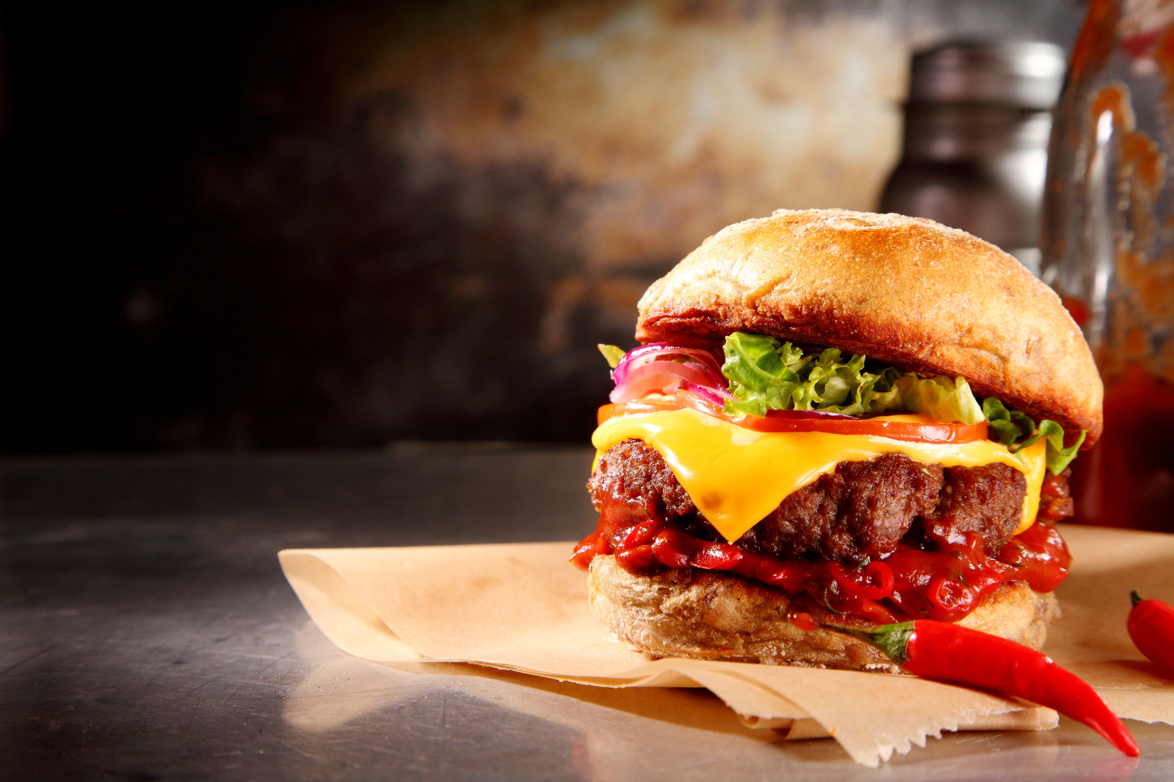 A burger with lettuce, tomato, and red sauce on a bun, placed on a table with a pepper grinder in the background.