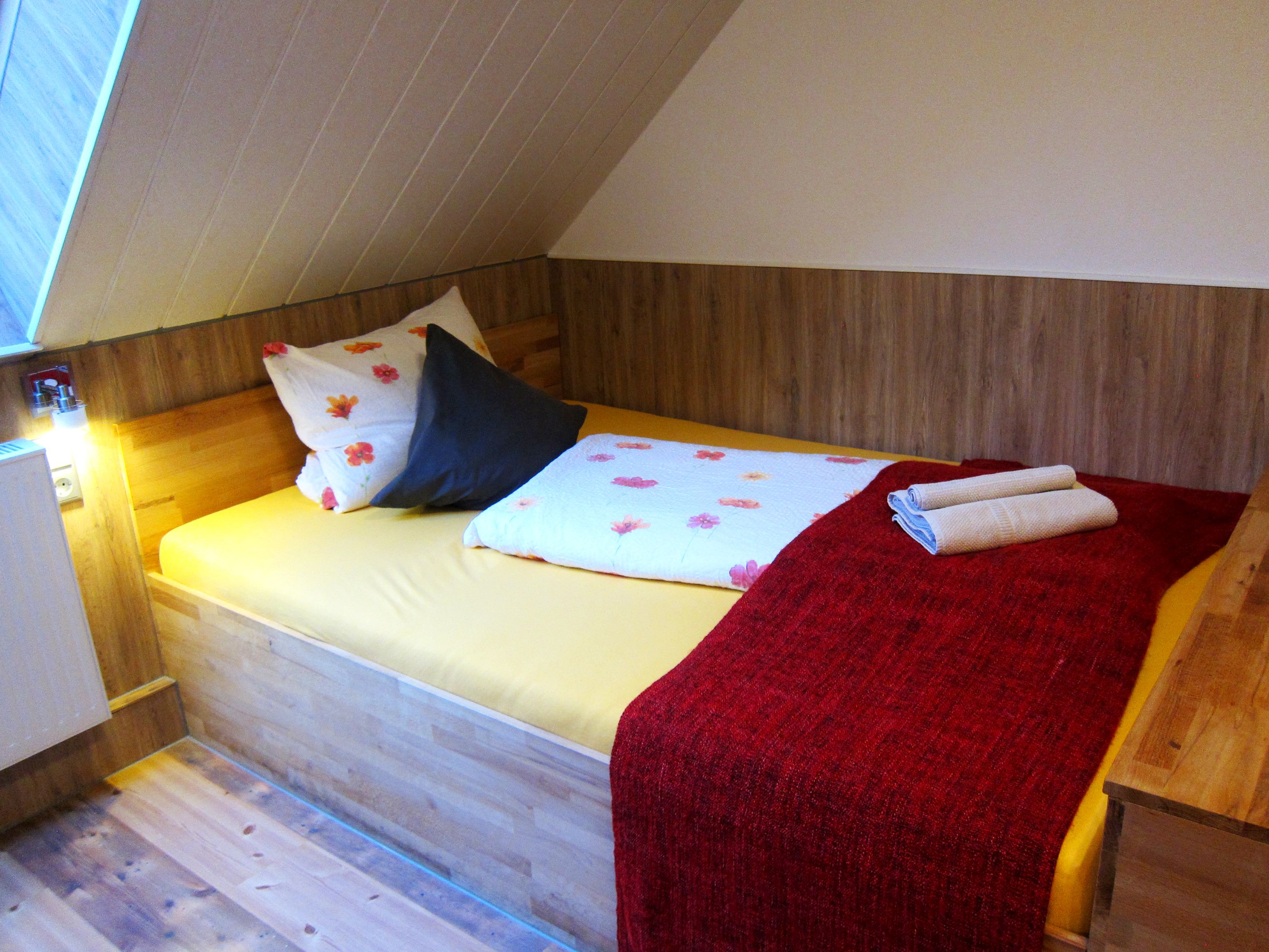 A small, cozy room with a wooden bed, a red blanket, and a pillow with a floral pattern.