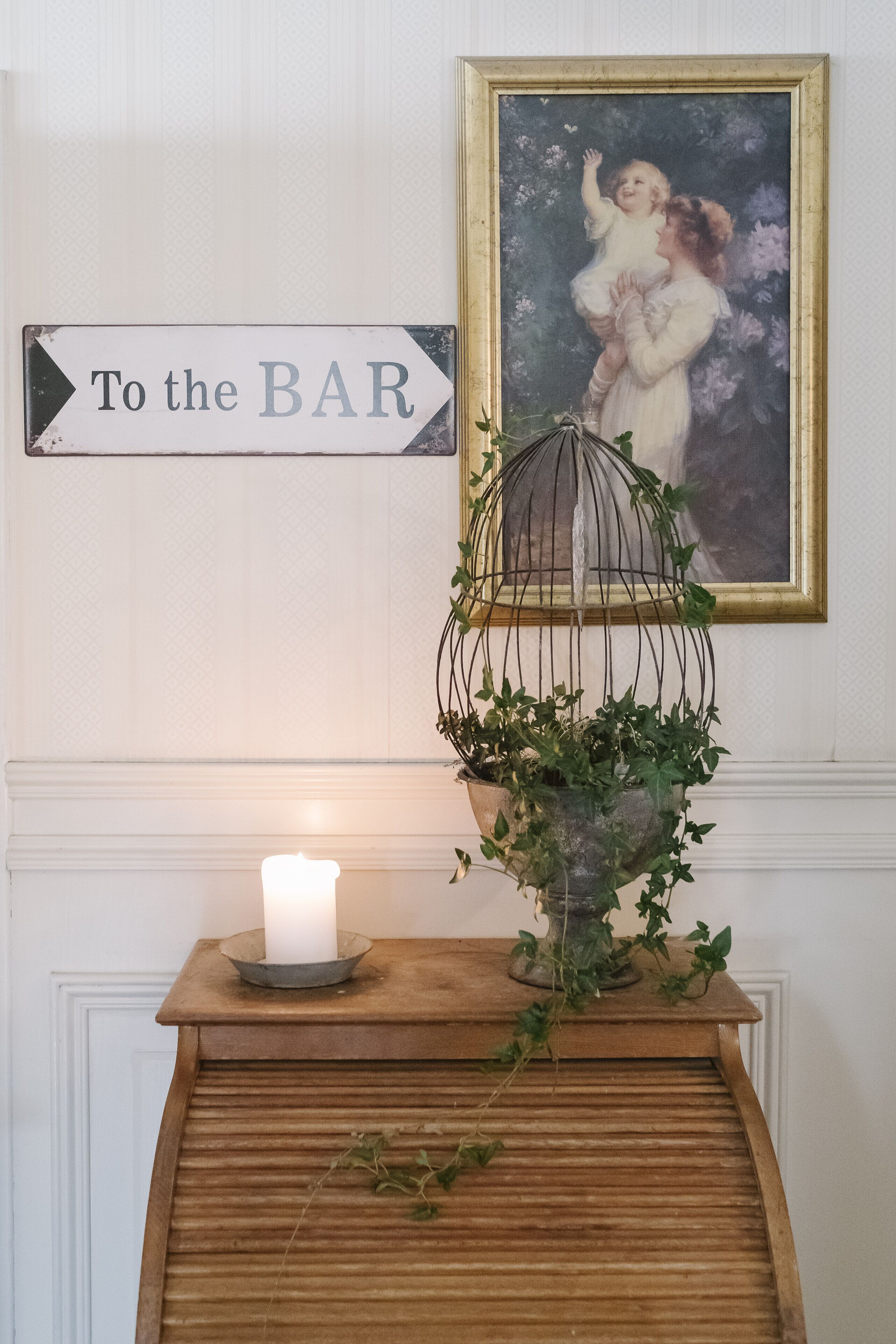 Une enseigne de bar, un tableau encadré, une bougie et une plante sur une table en bois.