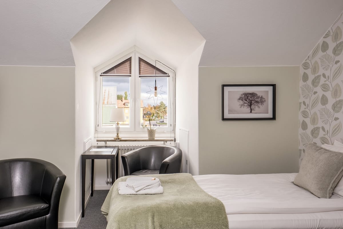 A hotel room with a bed, a desk with a chair, a window with a view of the outside, and a framed picture on the wall.