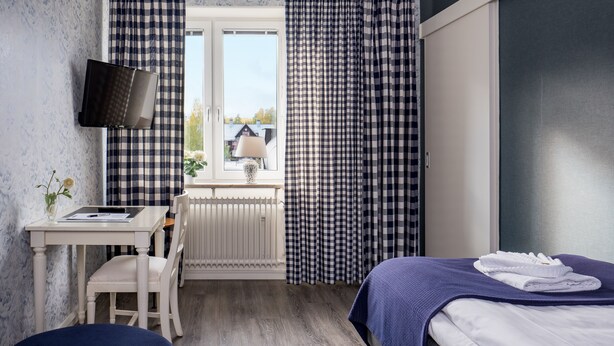 A hotel room with a checkered curtain, a bed with a blue blanket, a desk with a lamp, and a window with a view of buildings. - Hotel Vännäs (Vannas)