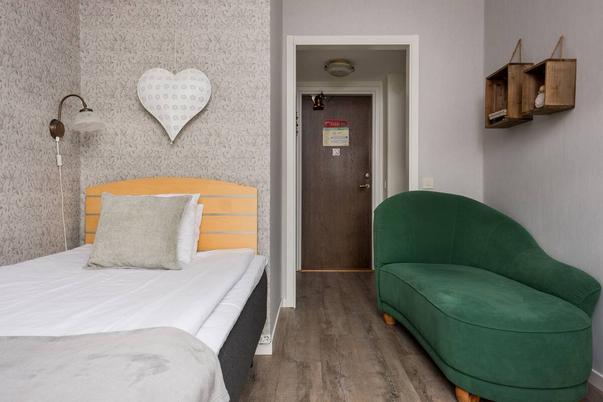 A room with a bed, a green sofa, a wall-mounted light fixture, and a door with a sign.