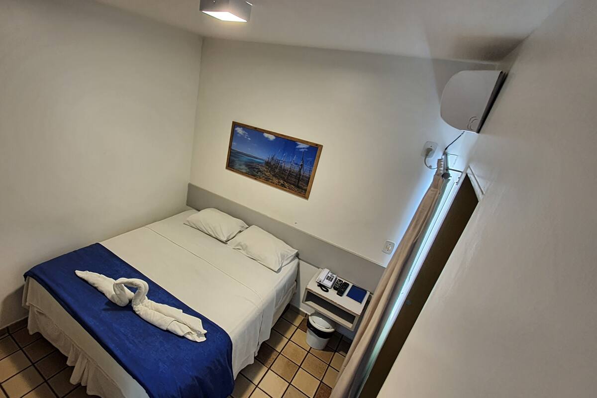 A hotel room with a single bed, a nightstand with a phone and a small table, a picture on the wall, and a bathroom with a toilet and sink.