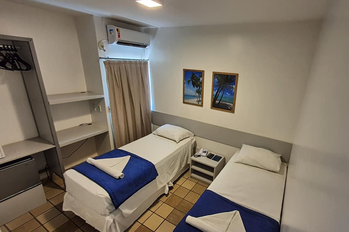 A hotel room with two beds, a desk, a wardrobe, an air conditioning unit, and two framed pictures on the wall.
