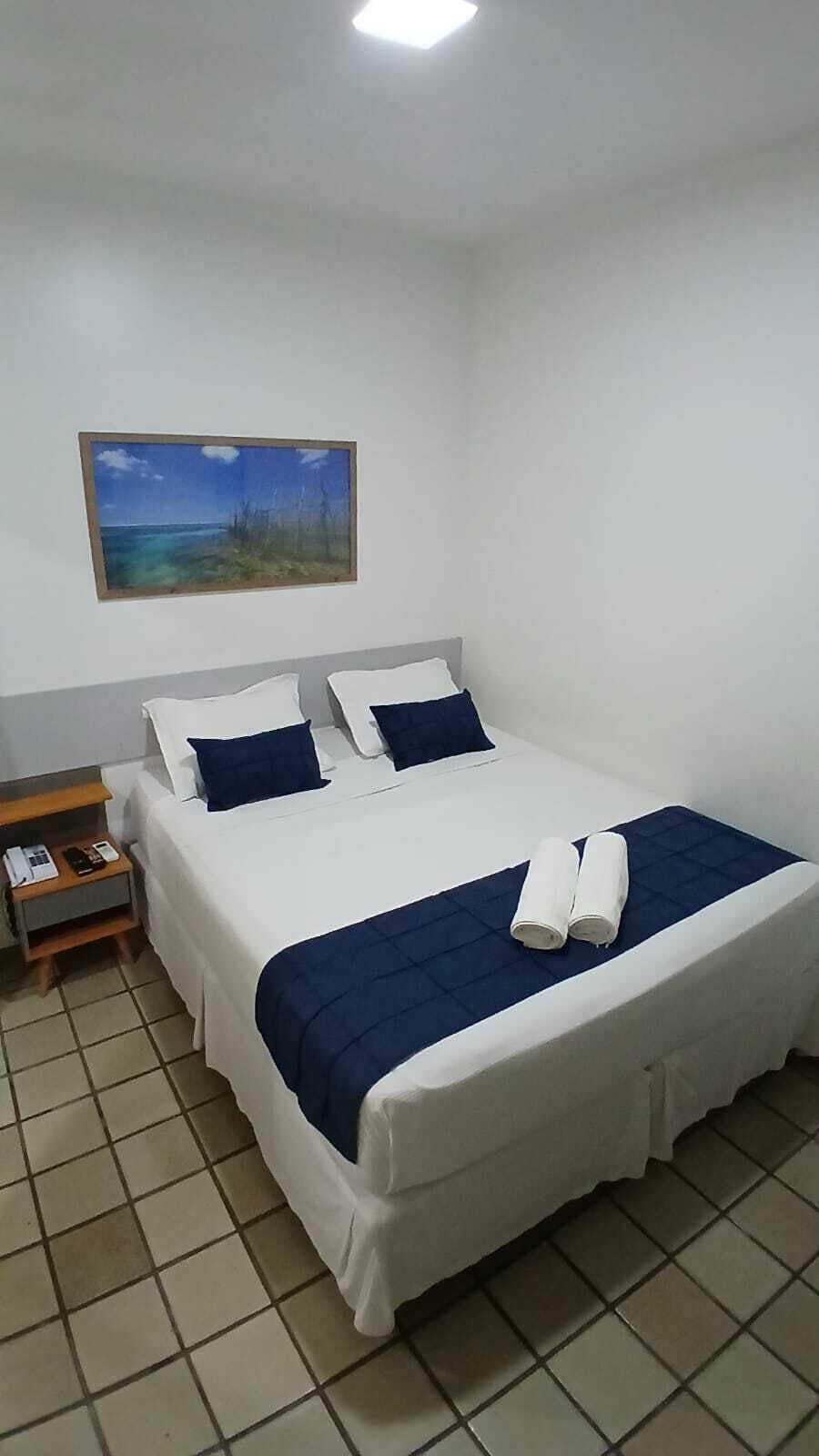 Ein Hotelzimmer mit einem Bett, einem Nachttisch, einem Gemälde und einem gefliesten Boden.