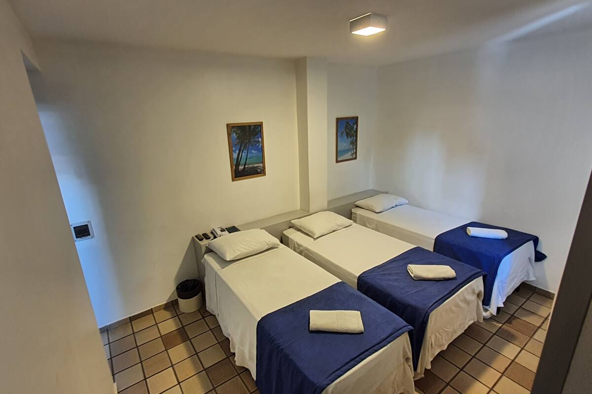 A hotel room with three single beds, a small bedside table with a lamp, and a trash bin.