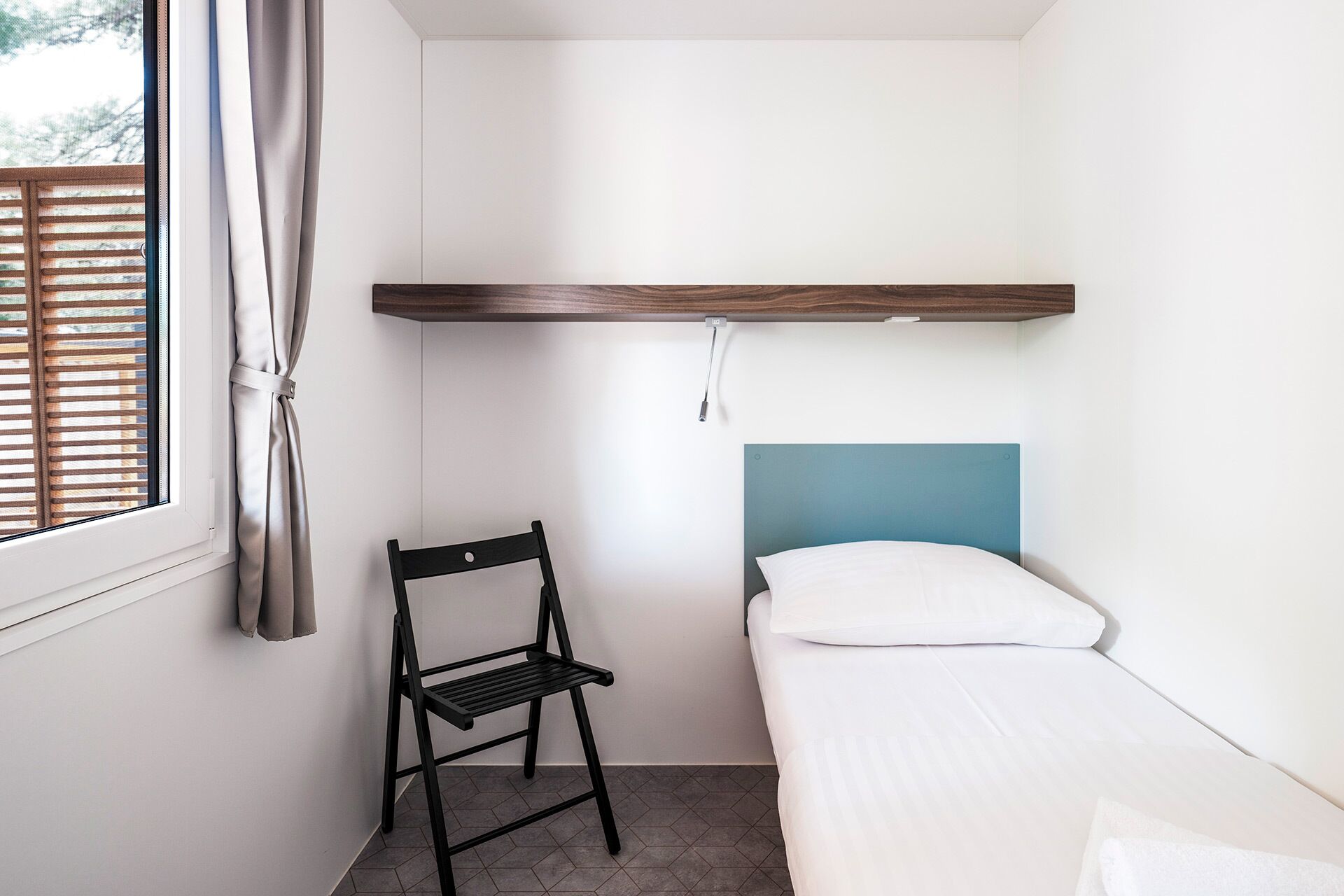 A small, single-bed room with a wooden shelf above the bed, a chair, and a window with blinds.