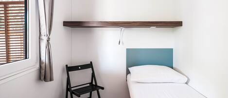 Una habitación pequeña con una cama individual, un estante de madera sobre la cama, una silla y una ventana con persianas.