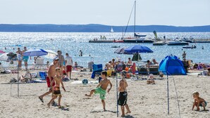 Una playa con gente jugando al fútbol, tiendas de campaña y barcos al fondo.
