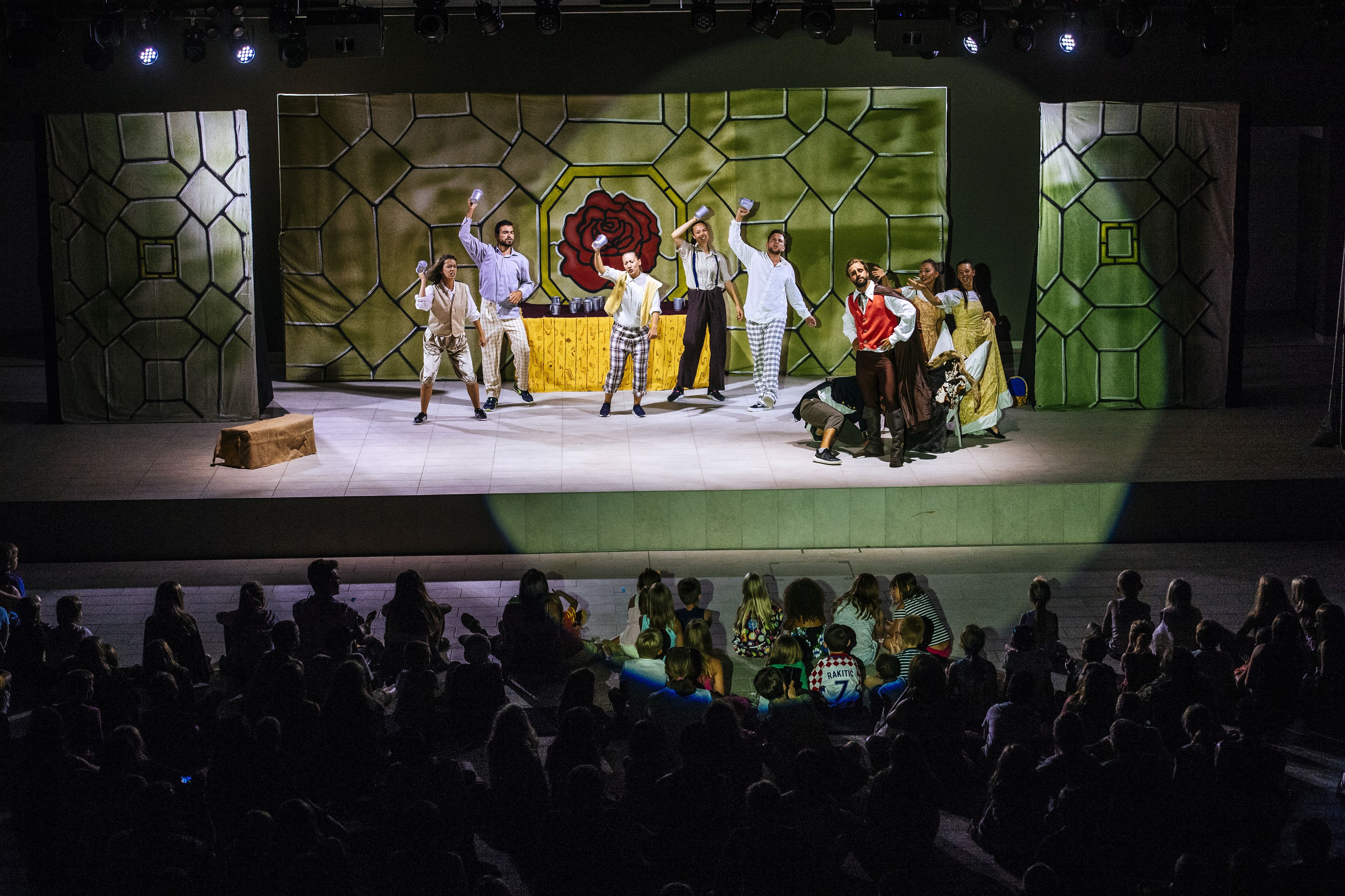 A stage with a group of people performing, a large audience seated in front, and a backdrop with geometric patterns and a large flower.