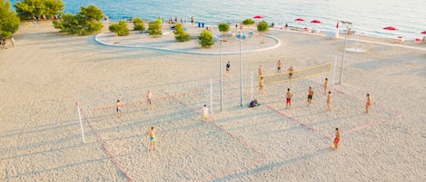A beach volleyball court with players and spectators, surrounded by trees and beach umbrellas.