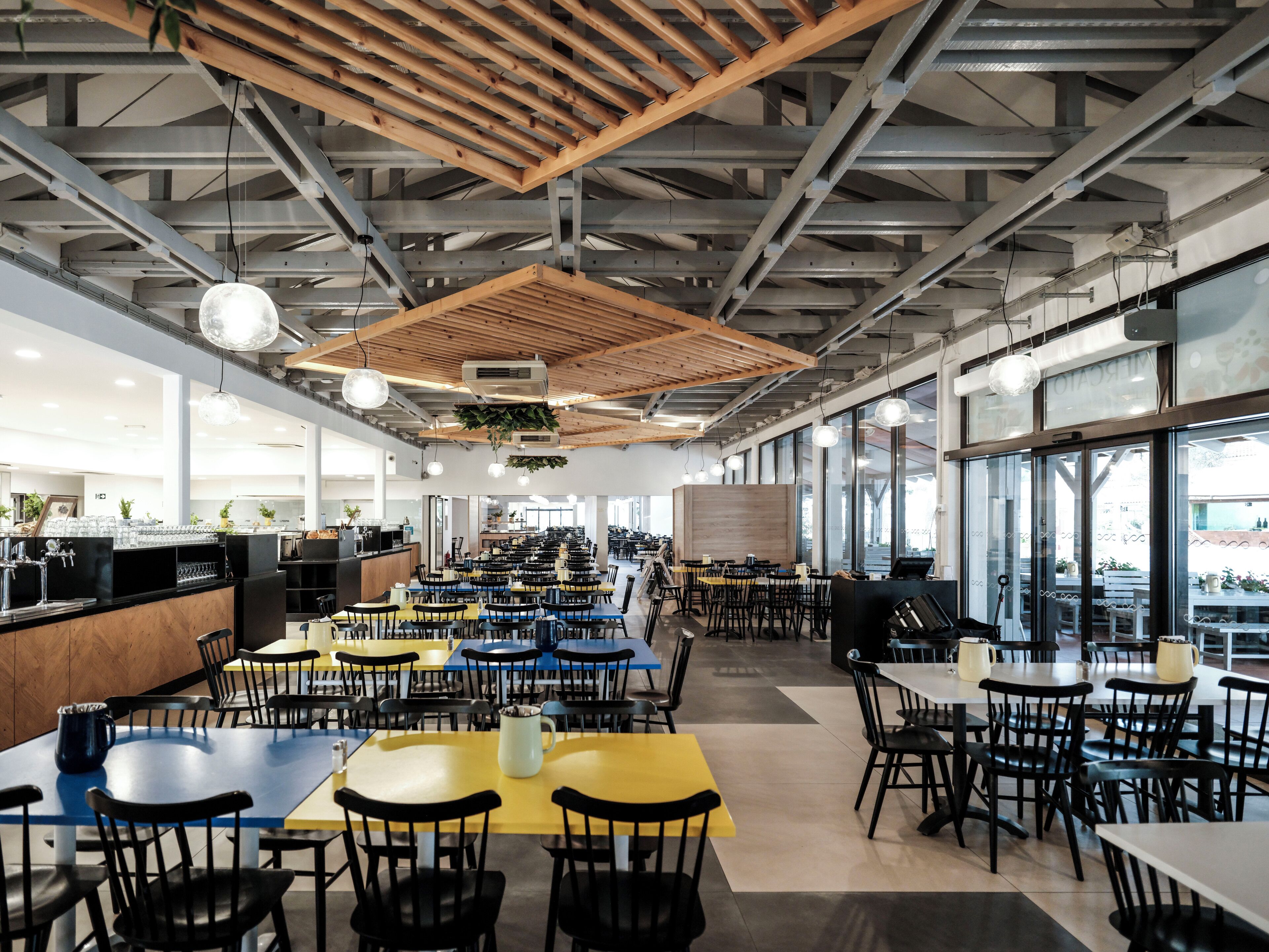 Eine geräumige Cafeteria mit mehreren Tischen und Stühlen, einem Küchenbereich und großen Fenstern, die für natürliches Licht sorgen.