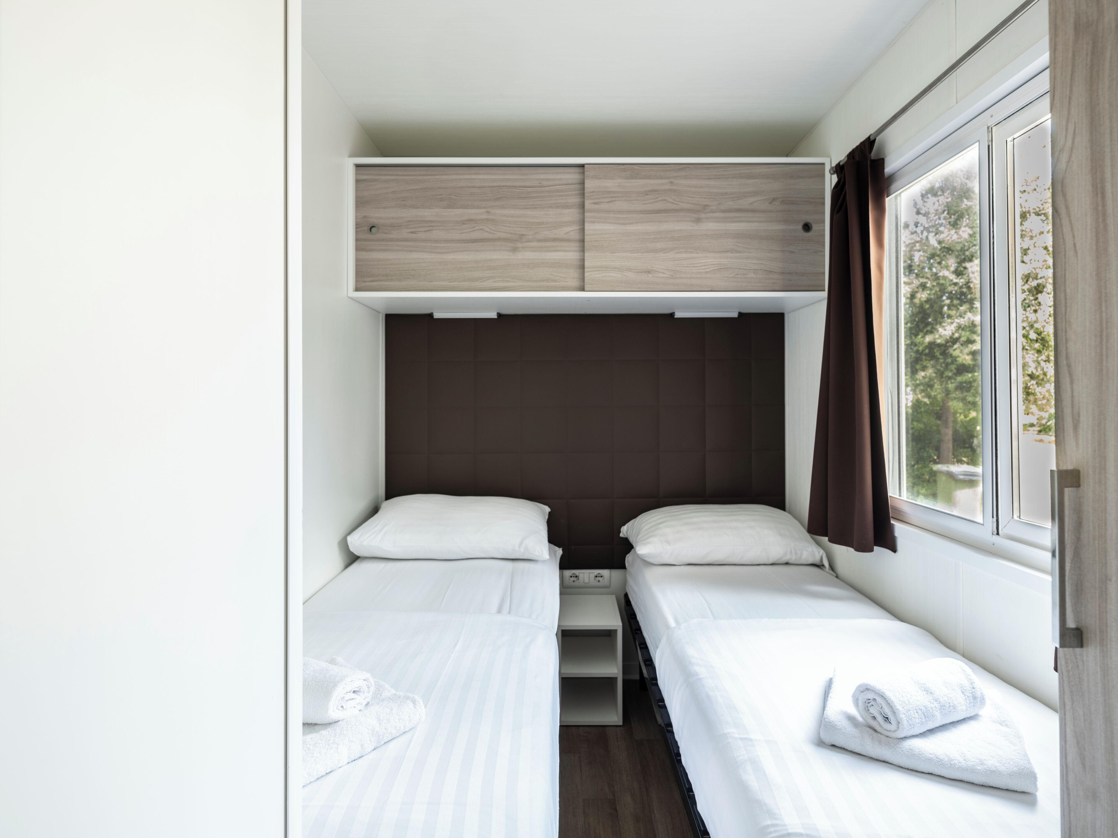 A compact bedroom with two single beds, a small storage unit, and a window with curtains.