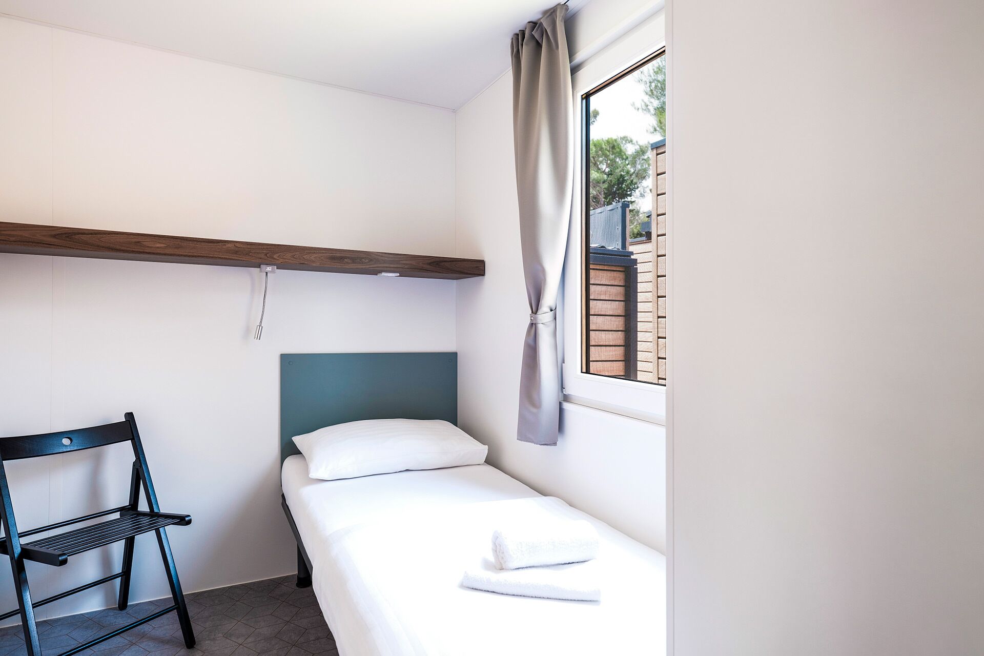 A single bed with a headboard, a wooden shelf above it, a chair, and a window with curtains.