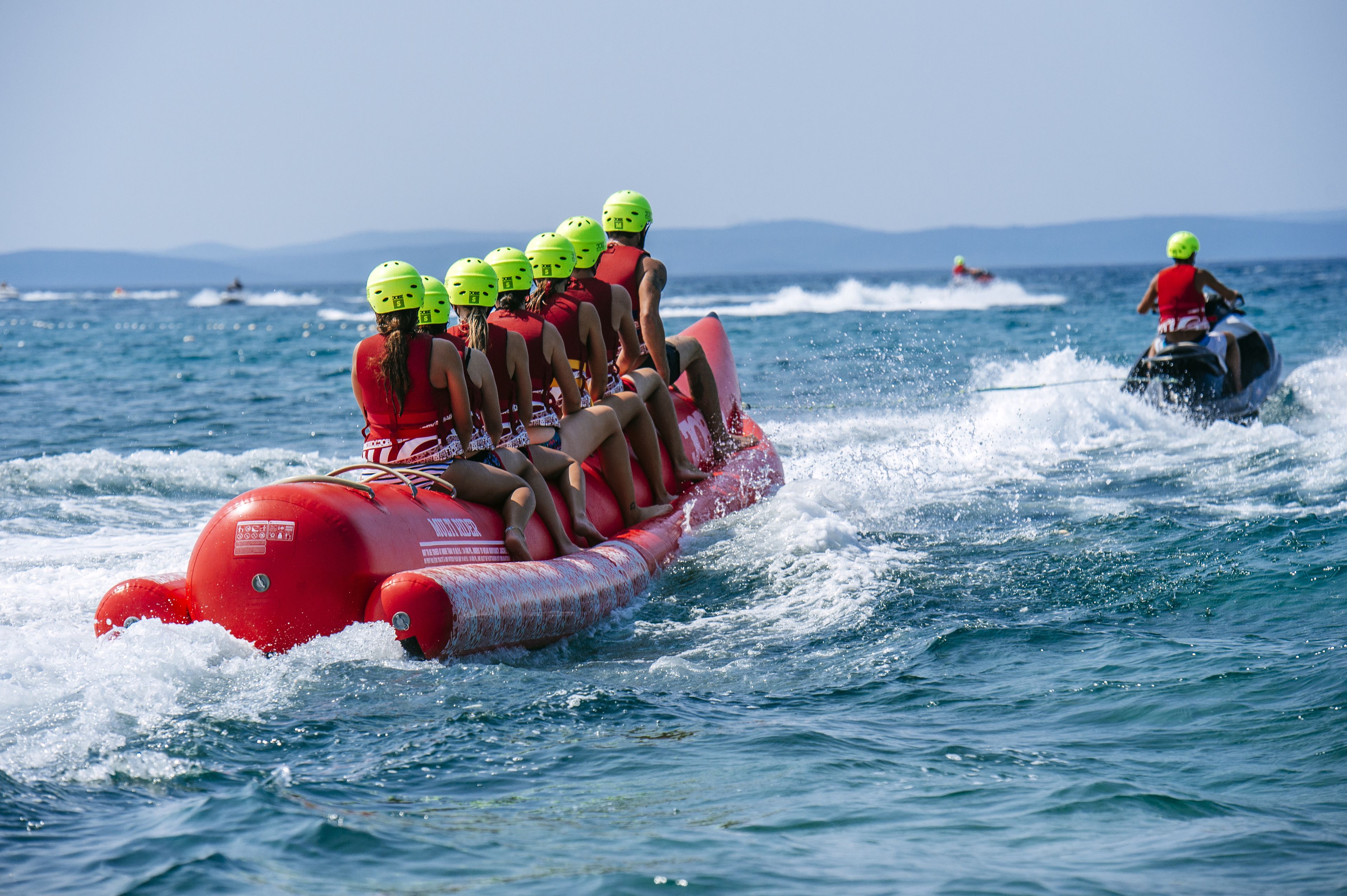 A group of people on a red inflatable raft with helmets, being towed by a jet ski.