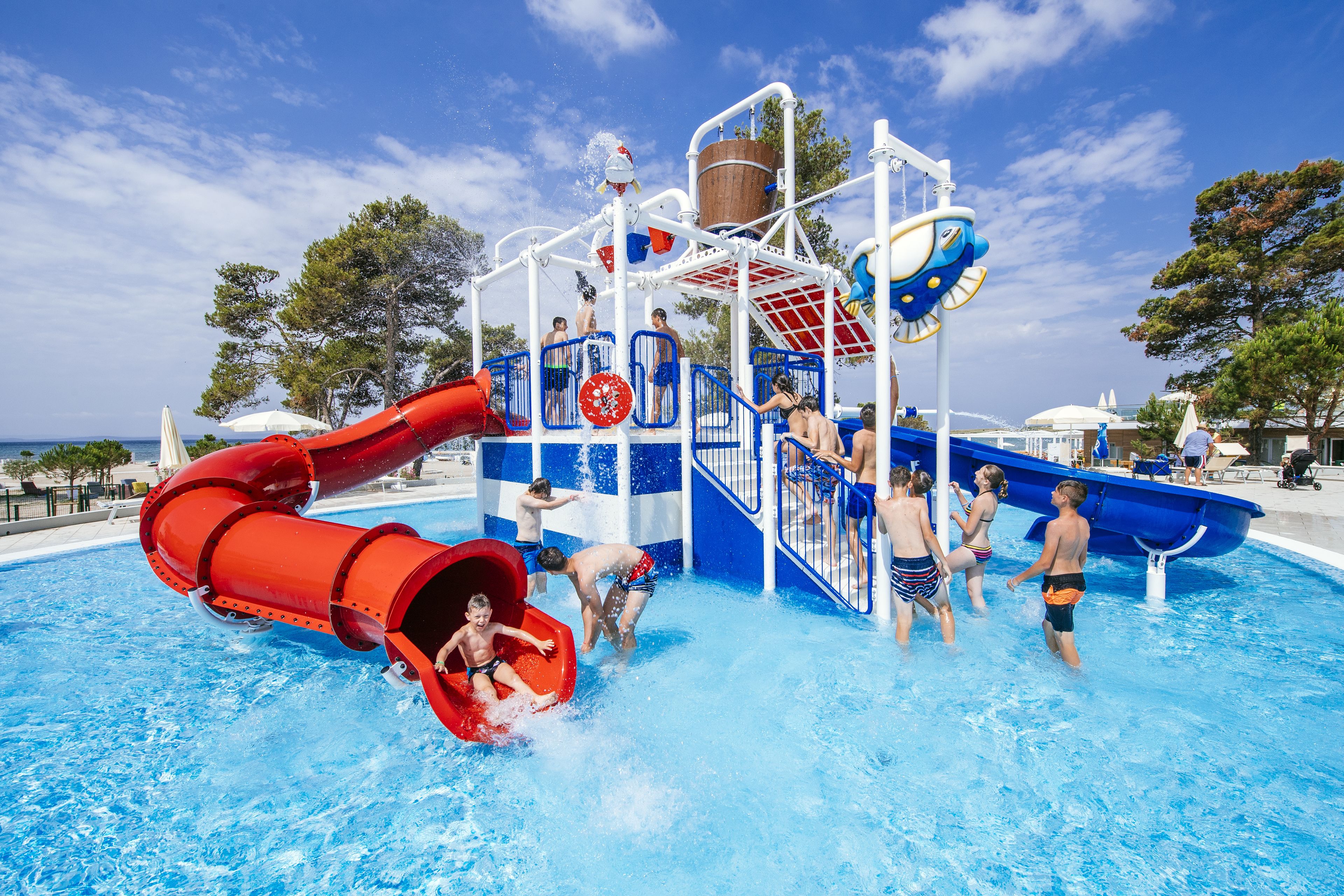 Parque acuático con un tobogán de agua rojo, zona de juegos acuáticos en azul y una piscina.