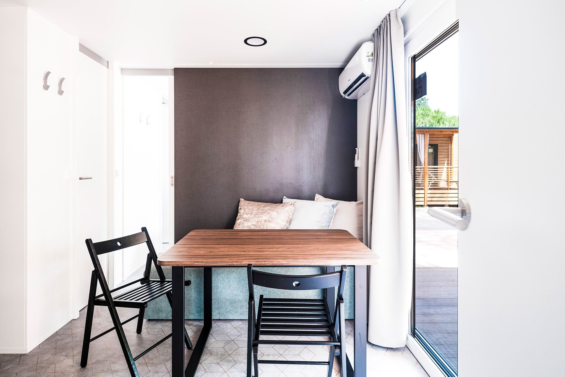A compact room with a wooden table and two chairs, a grey wall, and a window with curtains.