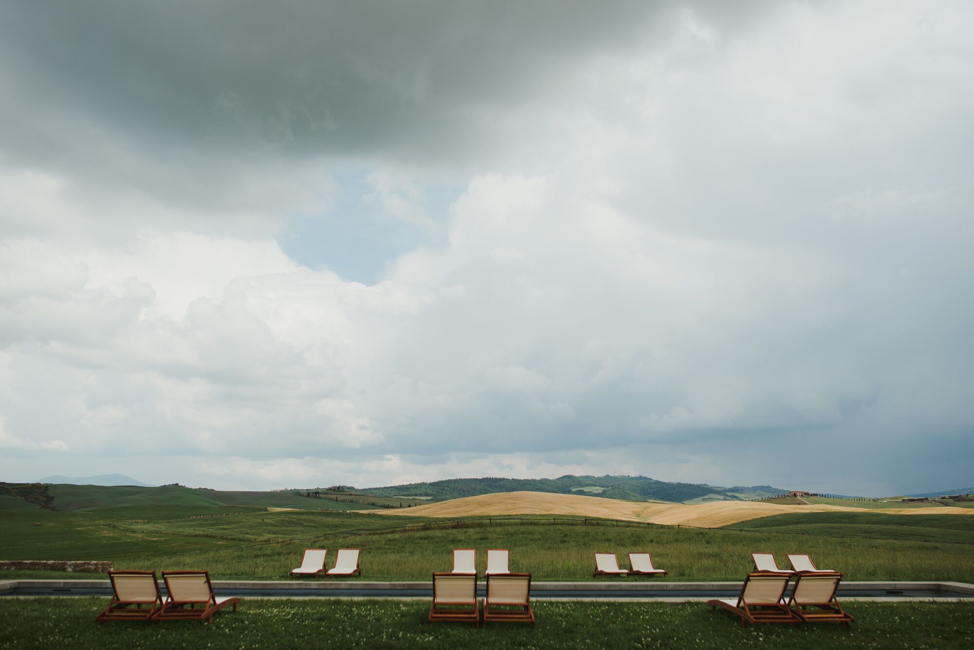 A row of empty lounge chairs facing a scenic landscape with rolling hills and a cloudy sky.