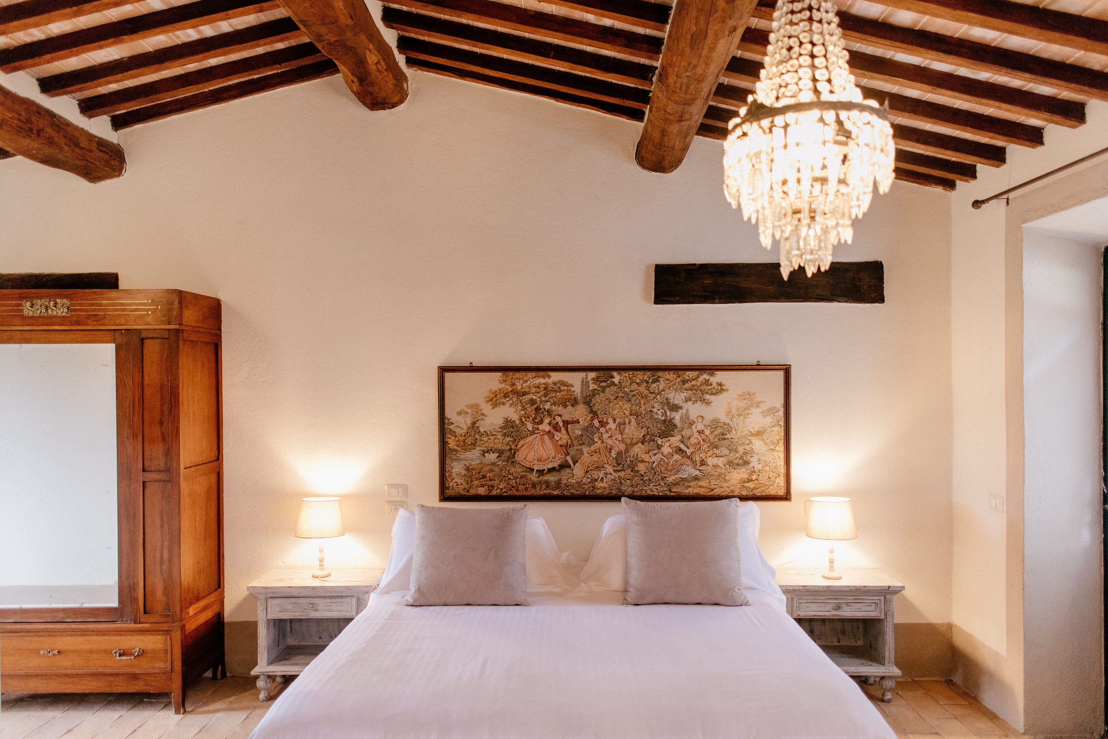 A bedroom with a large bed, wooden headboard, two bedside tables with lamps, a chandelier, and a large floral painting on the wall.