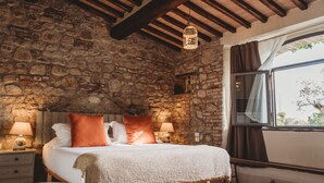 Une chambre aux murs de pierre, avec un lit, deux lampes de chevet, un banc en bois et une fenêtre avec des rideaux.
