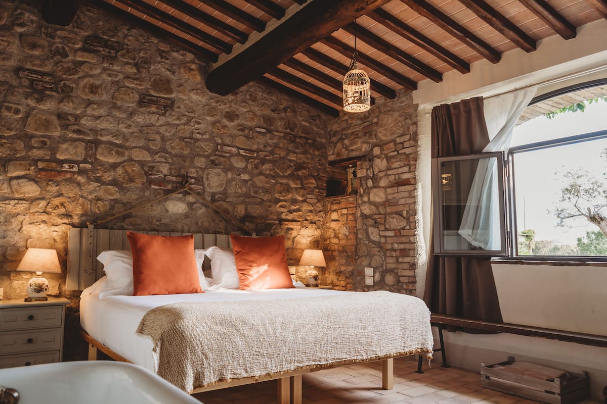 Ein Schlafzimmer mit Steinwänden, einem Bett, zwei Nachttischlampen, einer Holzbank und einem Fenster mit Vorhängen.