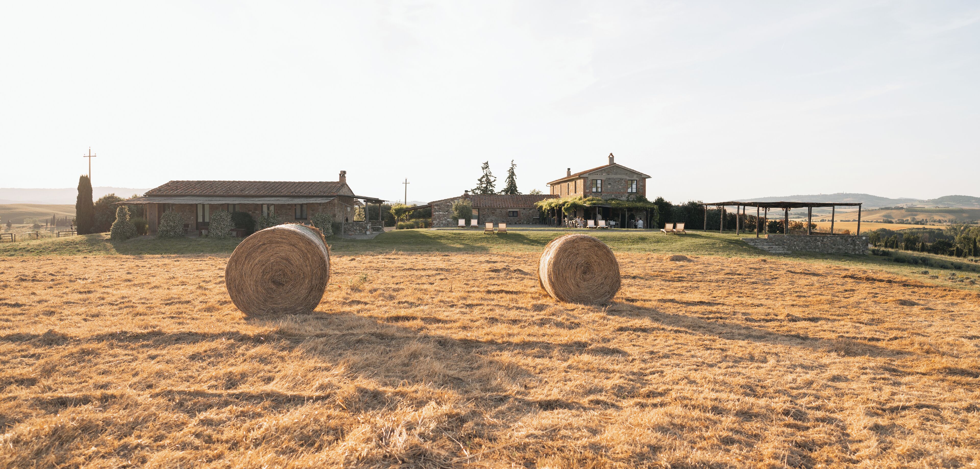 Un paysage rural avec deux bottes de foin dans un champ, un bâtiment en pierre et une maison plus grande dotée d’un patio couvert.