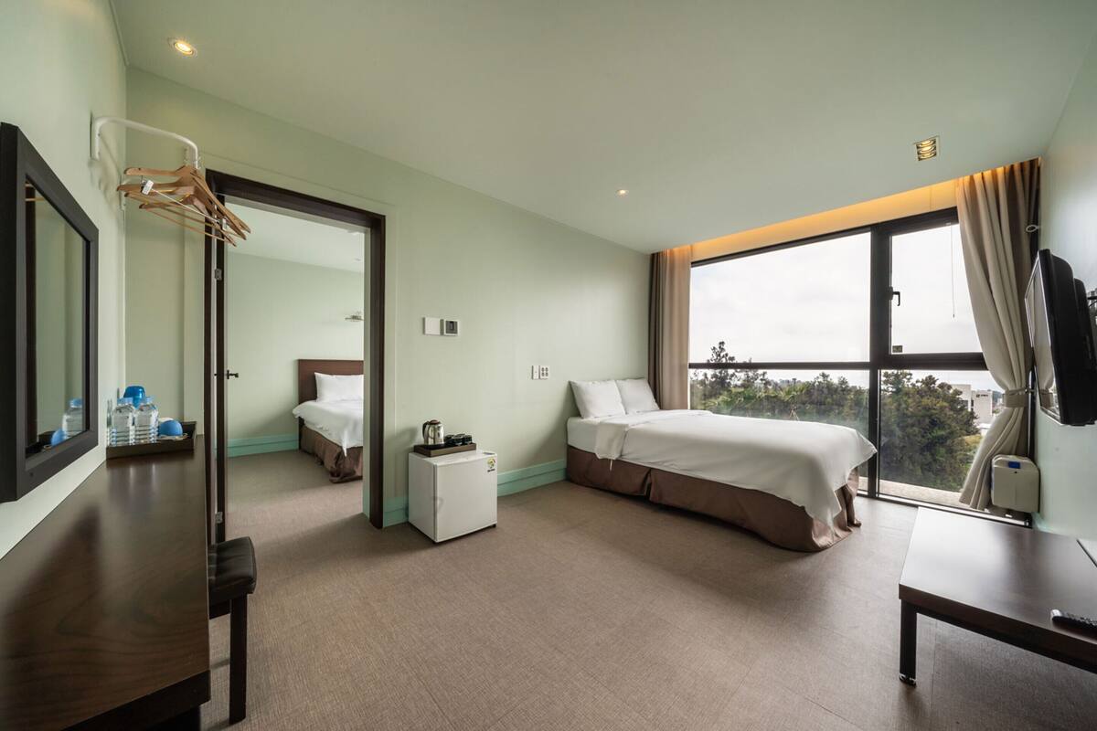 A hotel room with a bed, a desk, a small table, a mirror, a window with curtains, and a view of the outdoors.