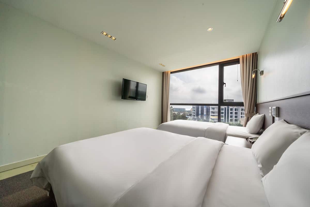A hotel room with two beds, a TV mounted on the wall, and a large window offering a city view.