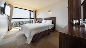 A hotel room with a large bed, a desk with a kettle, a flat-screen TV, and a city view. - Sunrise Hotel Jungmun (Seogwipo)