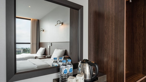 A hotel room with a large mirror, a bed with white linens, a bedside table with water bottles, a kettle, and a tray with toiletries. - Sunrise Hotel Jungmun (Seogwipo)