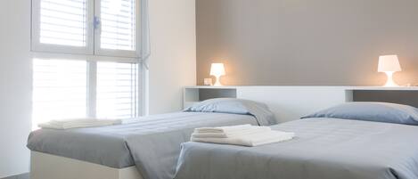 A hotel room with two beds, a window with blinds, a wall-mounted light, and a bedside table with a lamp.