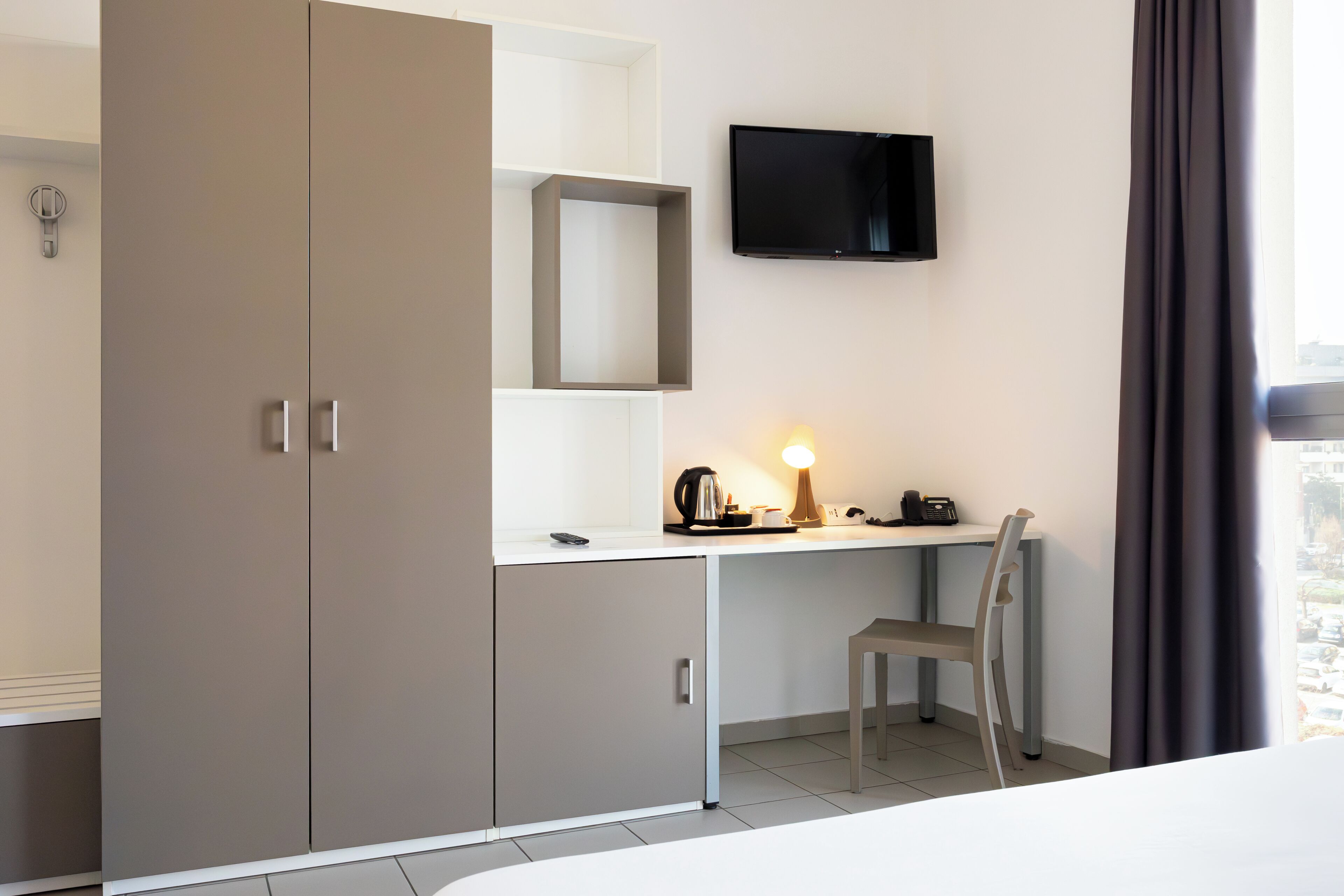 Ett modernt hotellrum med en stor garderob, ett skrivbord med en lampa, en stol och en tv som är monterad på väggen.