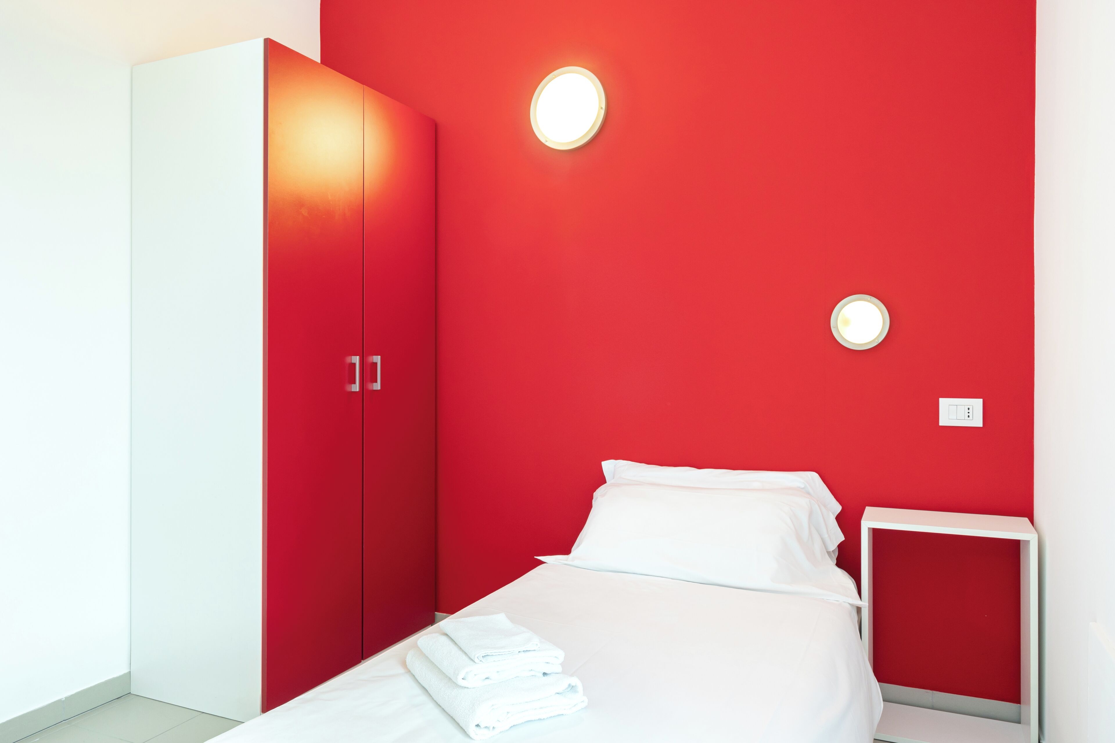 Una habitación de hotel moderna con una pared roja de acento, una cama blanca y un armario.
