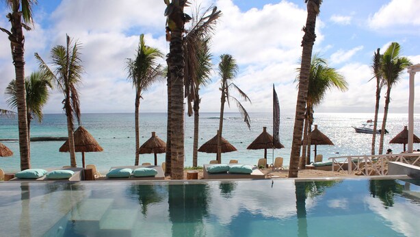 Zona de piscina con sillones de descanso y sombrillas de paja, con vistas a una playa y al océano.