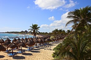 A beach with rows of thatched umbrellas and blue lounge chairs, palm trees, and a clear view of the ocean. - Bahia Principe Vacation Rentals - Four-Bedroom House (Akumal)