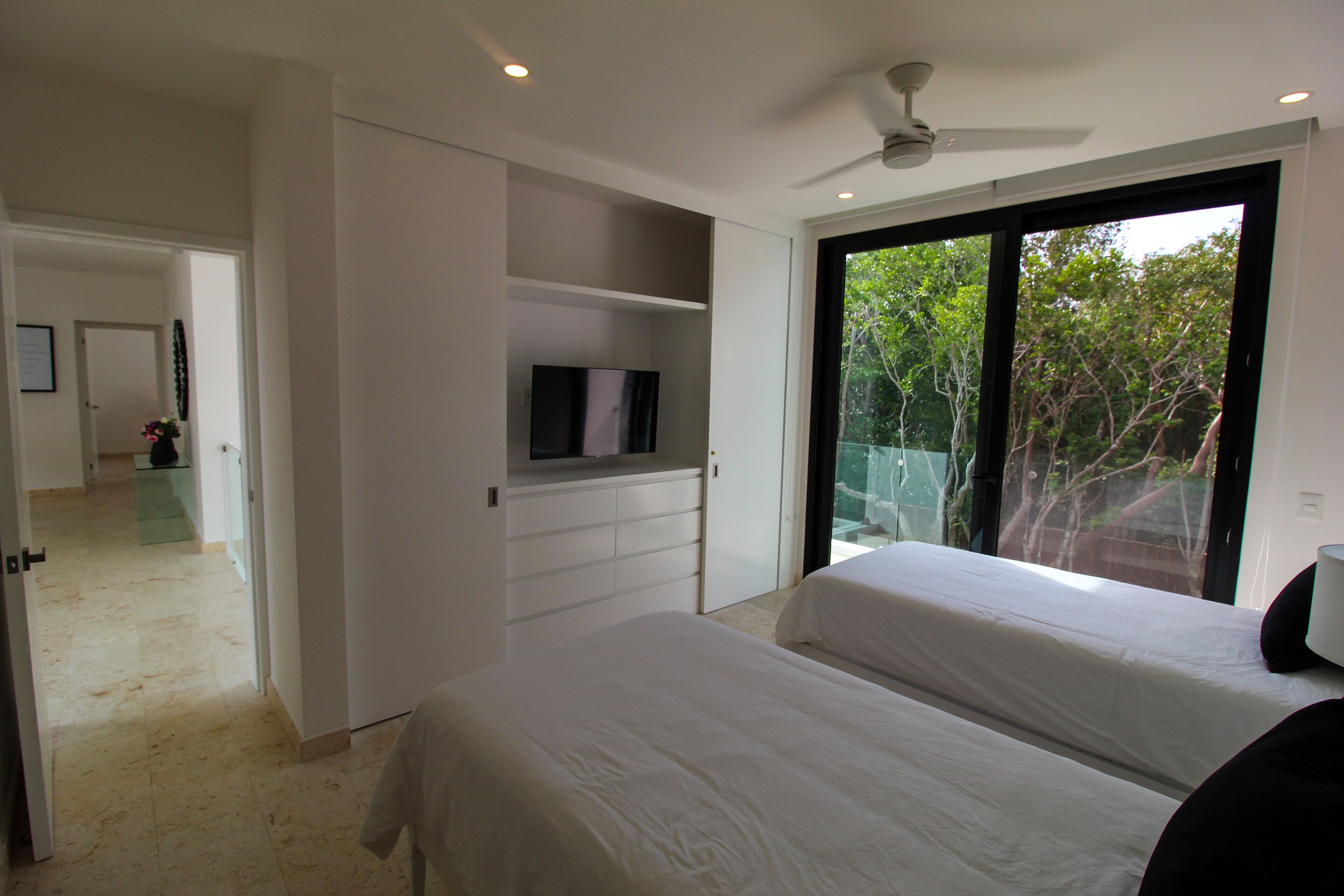 A bedroom with two beds, a TV on a shelf, and a large window with a view of trees.